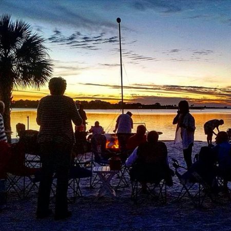 Groups of friends at sunset gathering around a campfire having drinks with friends on beach.